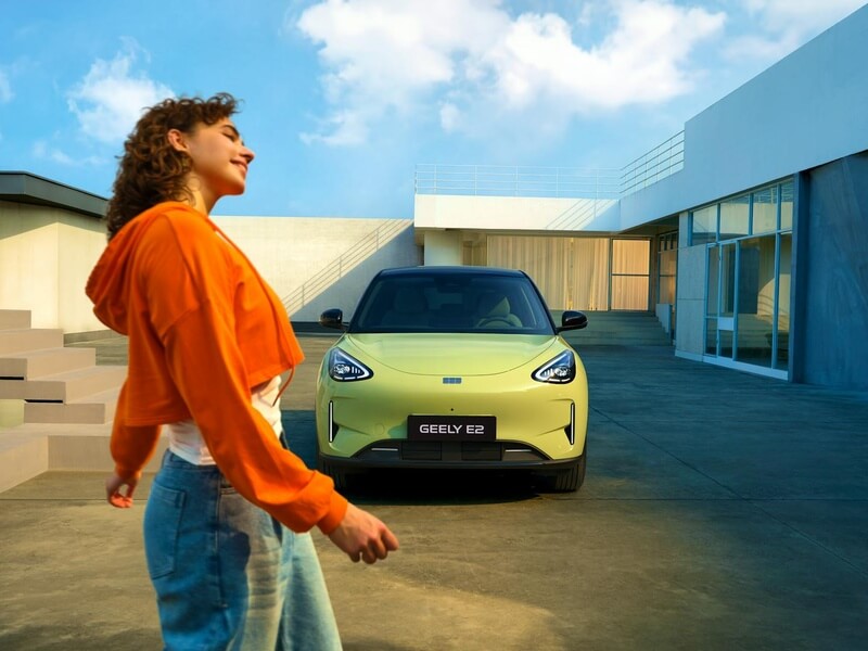 Geely E2 parcat în fața unei clădiri cu arhitectură modernă, cu o persoană zâmbitoare în prim-plan.